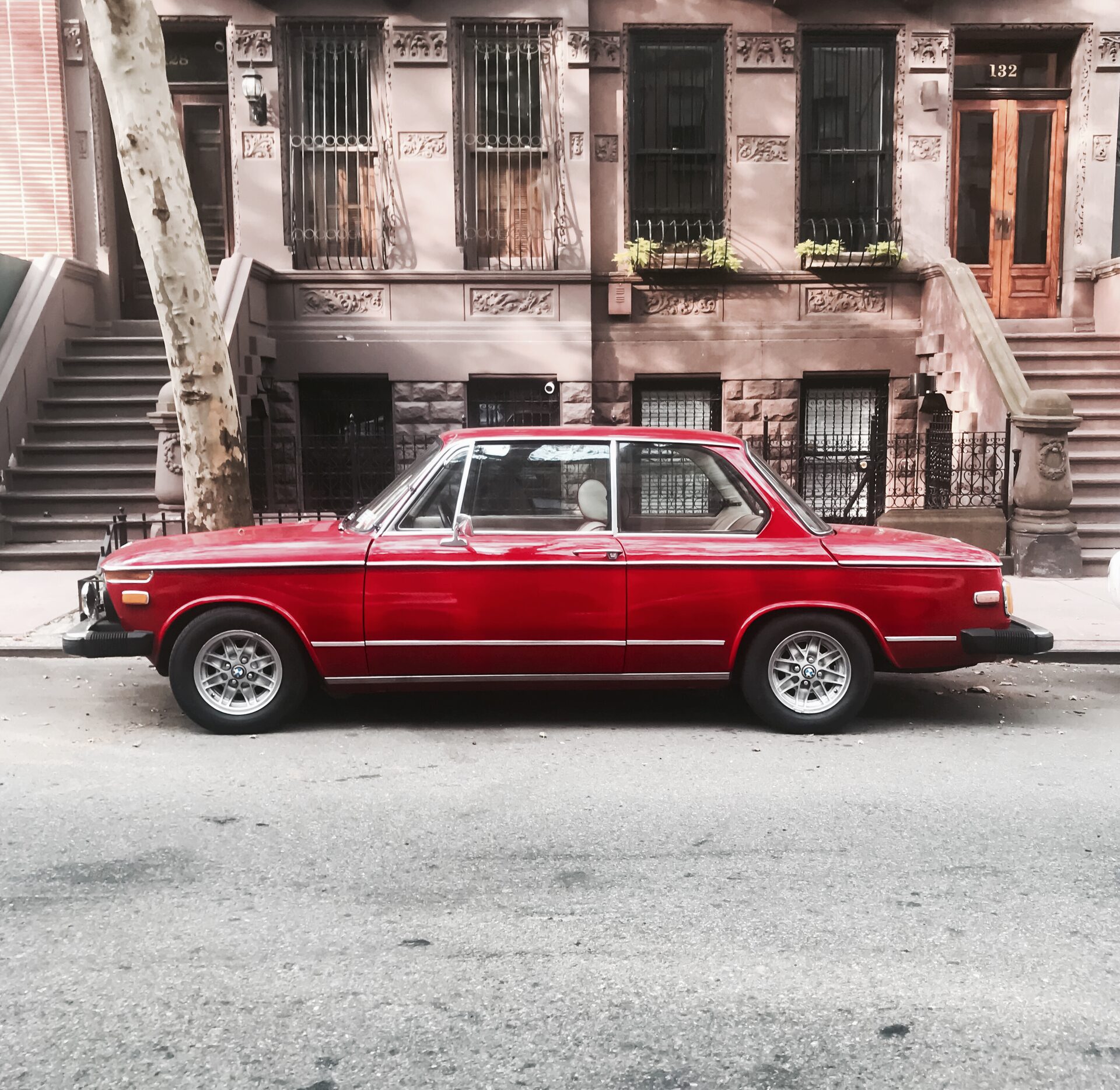 A red 1972 BMW 2002 parked on a tree-lined street