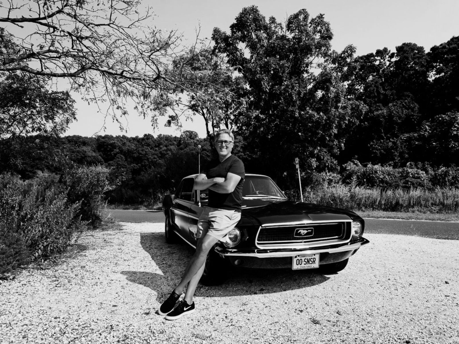 Tim Young leaning against his black 1968 Ford Mustang at a gravel pull-off, summer trees in the background — black and white.