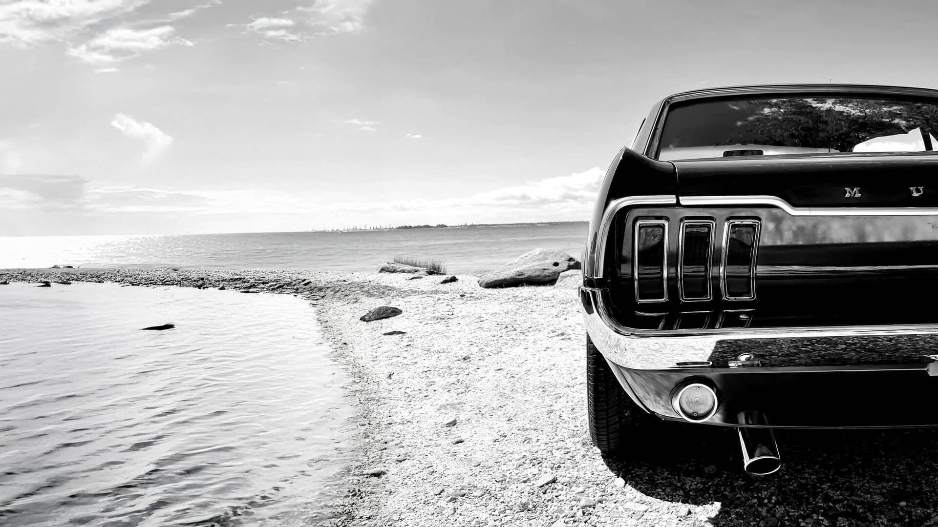 A 1968 Ford Mustang on a gravel beach, with water and distant shoreline — black and white.
