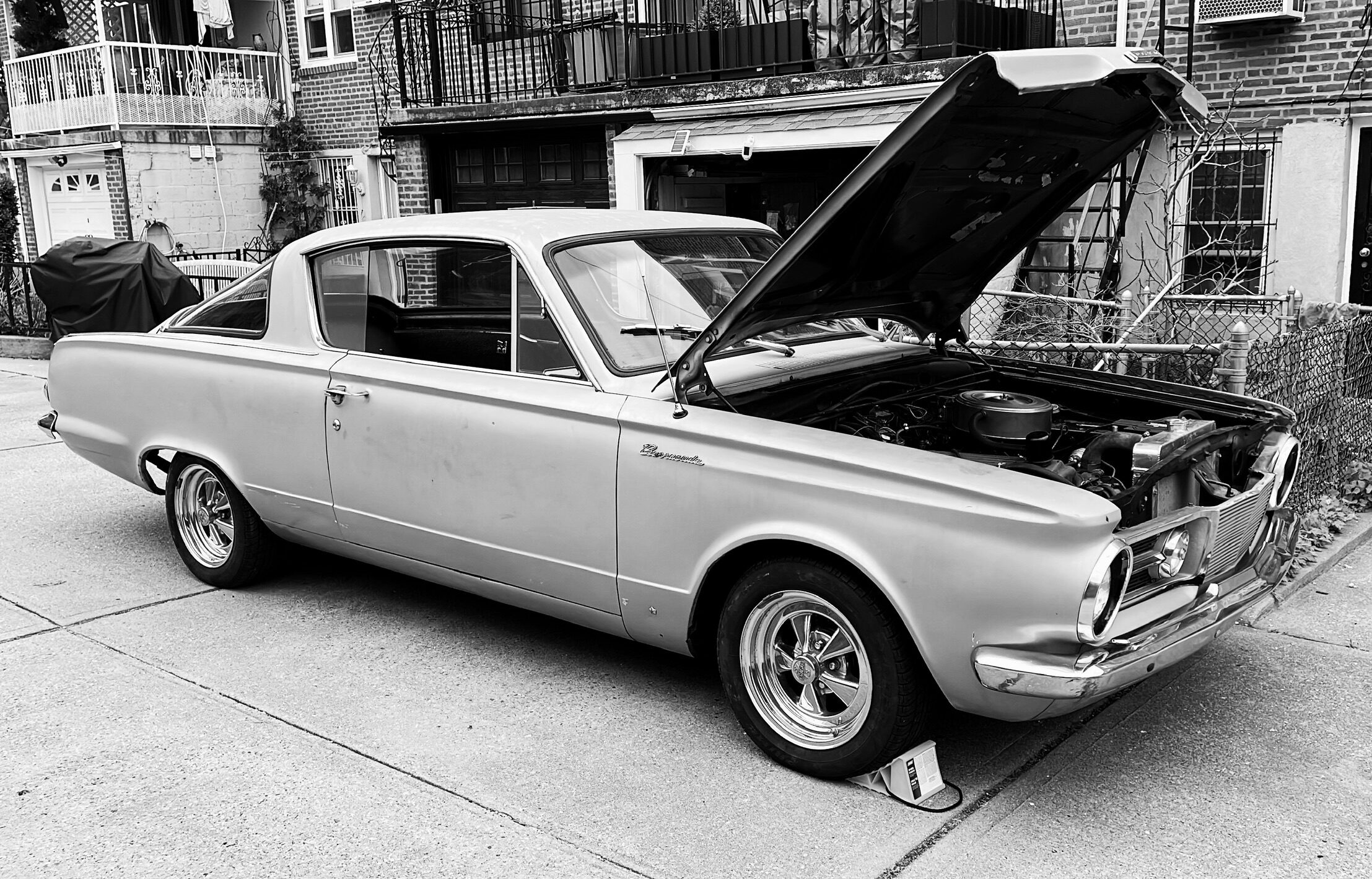 A silver 1964 Plymouth Barracuda parked on a driveway with its hood open, brick row-house garages behind — black and white.