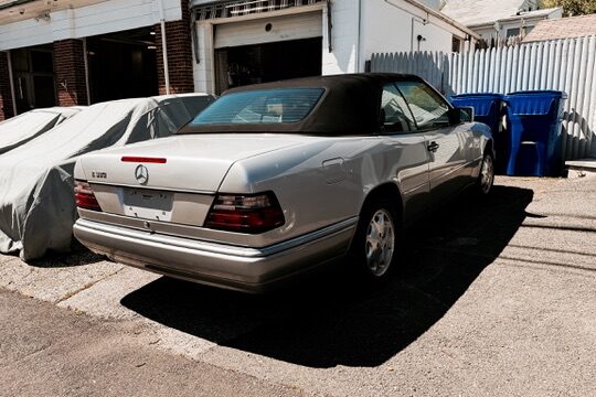 Silver 1995 Mercedes-Benz E320 Cabriolet, rear three-quarter right view with the soft top up, parked beside a garage