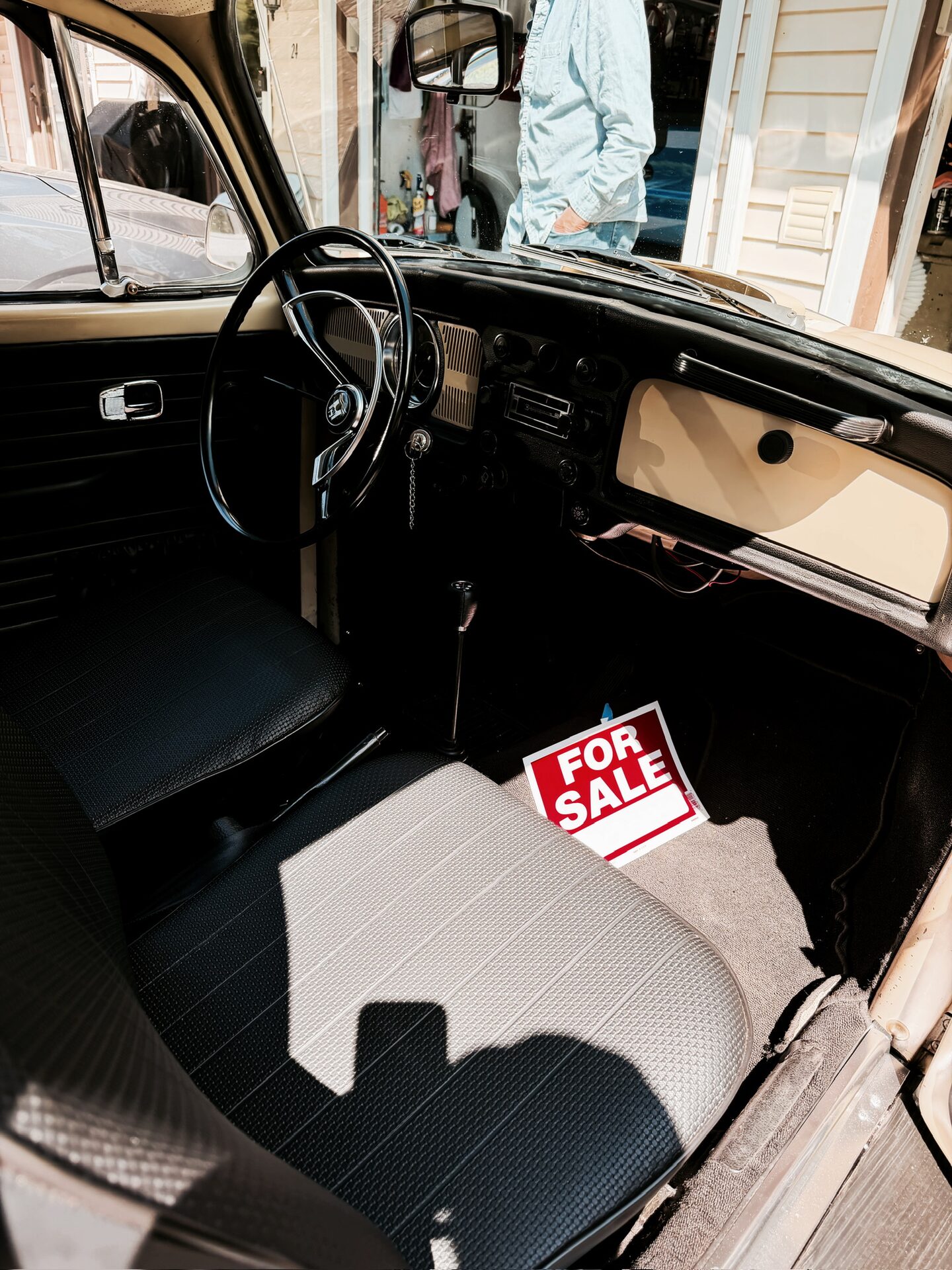 Interior of the 1969 VW Beetle &mdash; black dash, steering wheel, and bench seats with a For Sale sign on the passenger seat